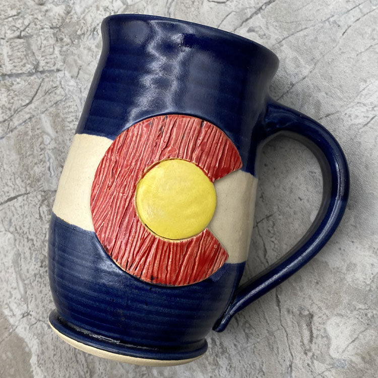 A dark blue handmade pottery coffee mug with a handle. Front features a textured colorado flag design.